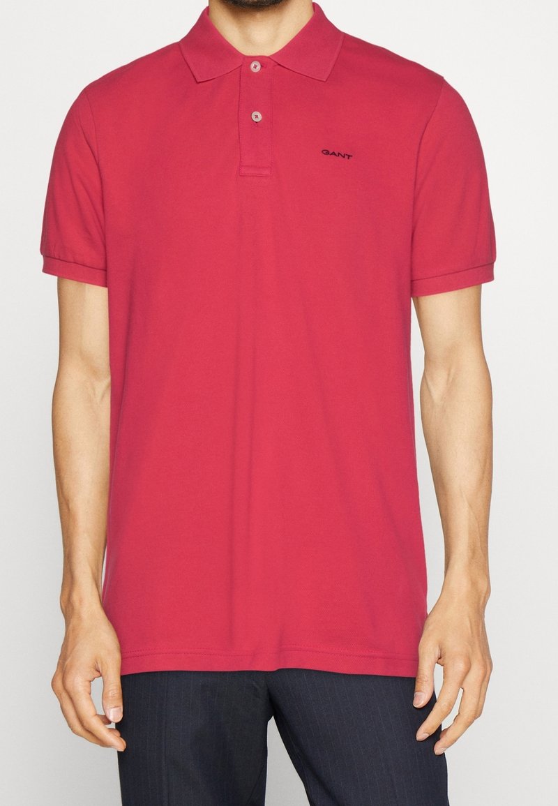 Homme portant un polo rouge à manches courtes avec deux boutons et un logo noir "GANT" sur le côté gauche de la poitrine, assorti à un pantalon foncé.