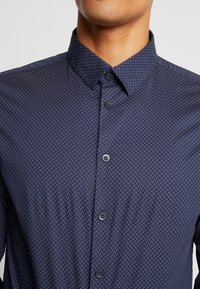 Marinblå knapp-up skjorta med ett litet vitt prickmönster, klassisk krage och en mjuk yta. Har kontrasterande knappar.