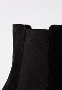 Acercamiento de una bota negra de ante hasta el tobillo con panel lateral elástico y costuras detalladas sobre un fondo gris claro.