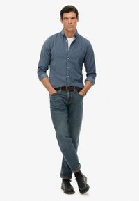 Uomo in piedi con sicurezza, con le mani in tasca, indossando una camicia blu con bottoni, maniche arrotolate, jeans blu, scarpe nere e un orologio al polso.