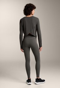 Nainen seisoo selkä kameraan päin, yllään hiilenharmaa pitkähihaisuus lyhyenmallinen paita, joka on päällekkäin takaa, sekä yhdistelevät legginsit ja mustat tennarit.