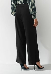 Lepas hitam lebar yang terbuat dari kain halus, dilengkapi dengan kantong belakang dan dipadukan dengan sandal hak tinggi hitam.