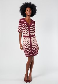 Lyhythihainen, vartalonmyötäinen mekko, jossa on V-pääntie. Mekossa on burgundinvärinen ja vaaleanpunainen siksak-kuvio sekä aaltoileva helma.