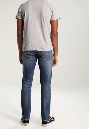 Mann steht mit dem Rücken zur Kamera, trägt ein graues T-Shirt, blaue Jeans und schwarz-weiße Turnschuhe vor einem einfarbigen Hintergrund.