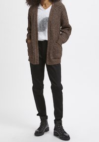 Personne portant un cardigan en maille marron, un t-shirt blanc à motif, un pantalon noir et des bottines noires à lacets, debout sur un fond blanc.