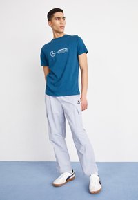 Teal t-shirt met witte "Mercedes AMG Petronas Formula One Team" logo, gecombineerd met lichtgrijze cargo pants en witte sneakers met zwarte accenten.