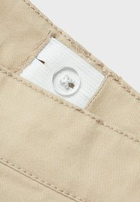 Tissu beige avec une étiquette blanche visible et un bouton transparent. Un détail de couture entoure l'étiquette près du bord supérieur. Matériau texturé.