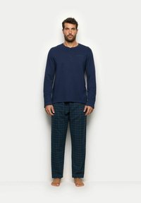 Marineblaues Langarm-Pyjama-Oberteil mit Knopfleiste, kombiniert mit grün karierter Hose. Aus Baumwolle, relaxte Passform, und barfüßige Füße sichtbar.