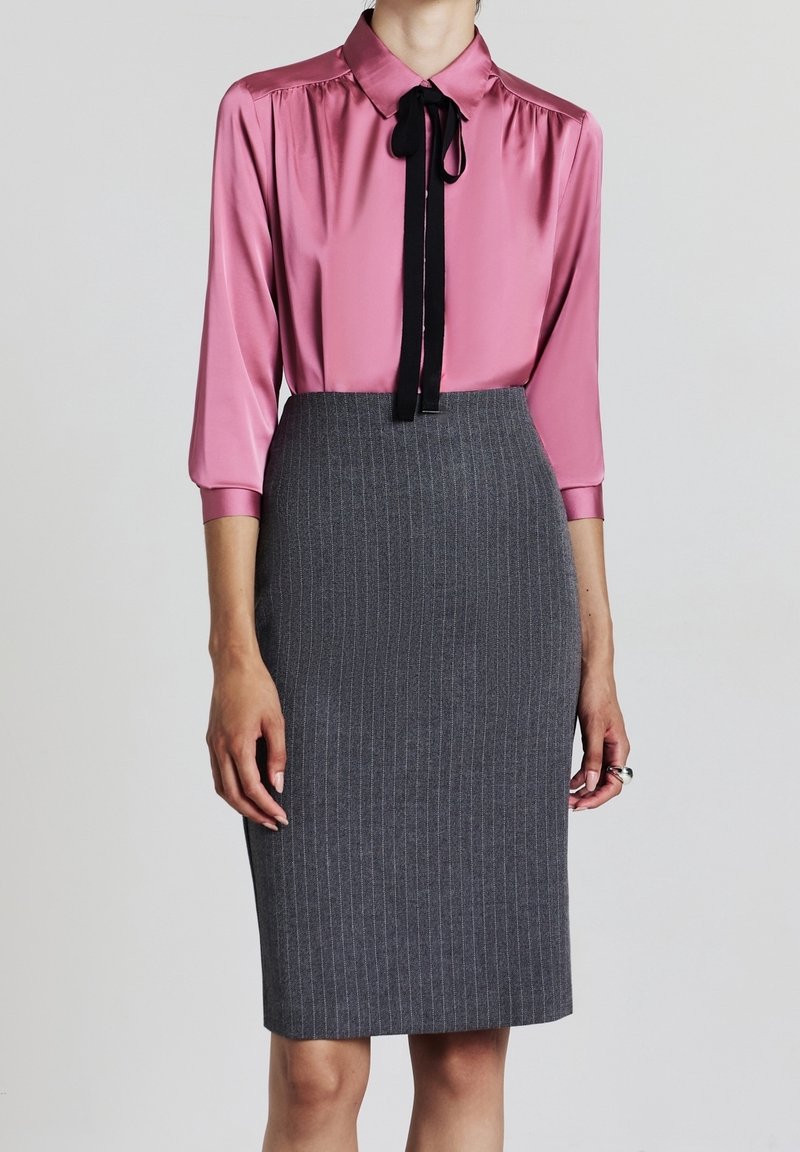 Femme portant un chemisier rose en satin avec une cravate noire et une jupe crayon grise à taille haute à rayures fines, debout devant un fond uni.
