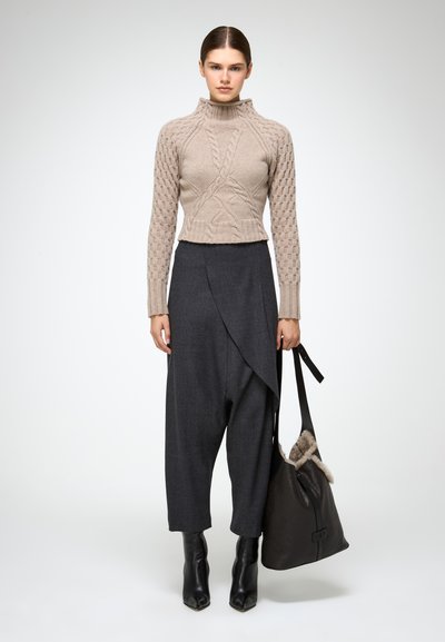 Mujer con suéter de cuello vuelto tejido en beige, pantalones de pierna ancha asimétricos en gris oscuro, botas de tobillo negras, sosteniendo un bolso de cuero negro con ribete de piel.