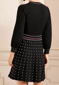 Černé šaty s přiléhavým vrškem a nafouknutými rukávy, texturovaným pasem a rozšířenou sukní zdobenou bílými čtvercovými vzory.