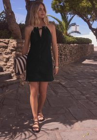 Robe noire sans manches avec un détail noué au niveau du cou, forme simple, portée avec des sandales noires ; sac rayé tenu dans la main gauche.