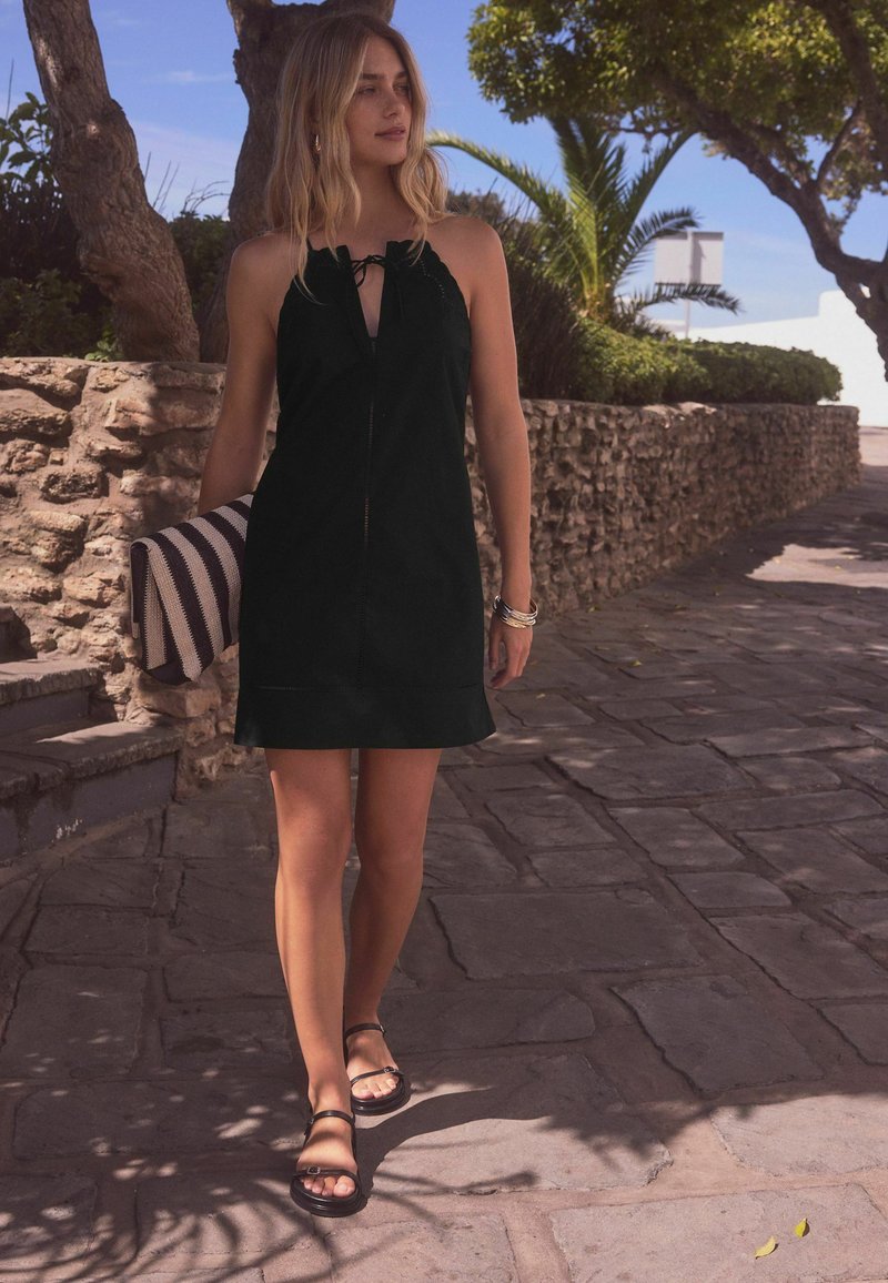 Robe noire sans manches avec un détail noué au niveau du cou, forme simple, portée avec des sandales noires ; sac rayé tenu dans la main gauche.