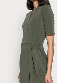 Olivgrön omslagsklänning i mjukt, texturerat tyg med korta ärmar, en knytbälte i midjan och en V-ringad halslinning.
