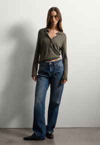 Camisa de botones de manga larga en verde oliva combinada con jeans de pierna ancha en azul oscuro y zapatos negros. Tela suave con ligero brillo, corte relajado.