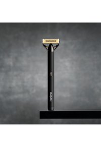 Elektrisk barbermaskin med en slank svart kropp, gullfarget metallblad og en knapp på siden. Plassert på en svart overflate.