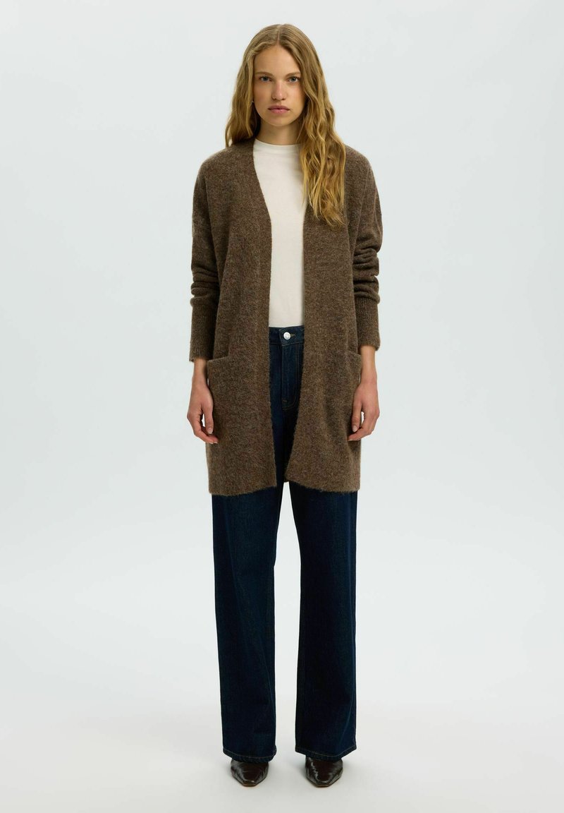 Femme portant un long cardigan marron sur un haut blanc, un jean large foncé et des chaussures pointues foncées, debout devant un fond uni.
