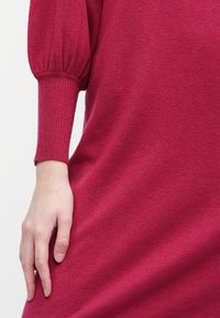 Suéter de malha em rosa brilhante, com um corte descontraído, punhos canelados e uma textura suave. Mão posicionada ao lado, mostrando os detalhes do tecido.