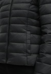 Μαύρο καπιτονέ μπουφάν με φερμουάρ στο μπροστινό μέρος και πλαϊνή τσέπη, που δείχνει τη μέση και το χέρι, φορεμένο πάνω από μαύρο παντελόνι.