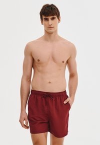 Czerwone kąpielówki z elastycznym pasem i sznurkiem, wykonane z lekkiego materiału. Bez widocznych wzorów ani akcentów.