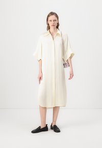 Kremkrāsas garš krekla kleita ar apkakli, īsām platām piedurknēm un taisnu siluetu, apvienota ar melniem slip-on apaviem.
