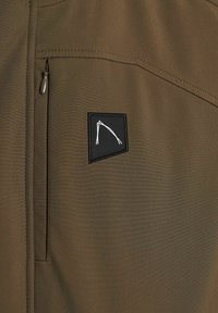 Veste vert olive avec une poche latérale, ornée d'un patch rectangulaire noir avec un contour de logo blanc. Texture de tissu lisse. Tirette de fermeture éclair visible.