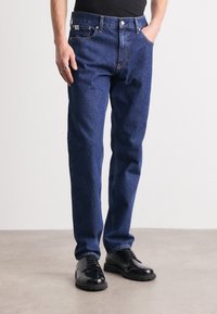 Tumedat sinised teksad klassikalise lõikega, viie taskuga ja peente õmblusdetailidega, koos mustade ametijalanõudega.