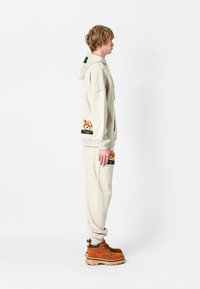 Sudadera beige y joggers a juego con parches de osito de peluche en los bolsillos. Botas marrones con acentos negros. Tela suave y lisa, corte relajado.