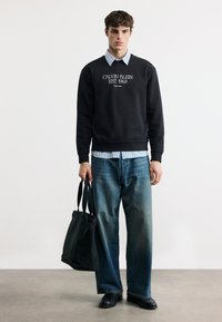 Νέος άνδρας με μαύρο φούτερ Calvin Klein, ριγέ πουκάμισο, φαρδιά μπλε τζιν, μαύρα παπούτσια, κρατώντας μαύρη τσάντα ώμου, στέκεται σε γκρι πάτωμα.