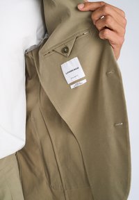 Χέρι που κρατά ανοιχτό ένα μπεζ σακάκι, δείχνοντας την εσωτερική τσέπη με κουμπί και λευκή ετικέτα που γράφει "LINDBERGH Regular Fit Superflex Recycled Fibers."