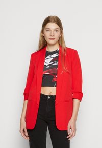Červený blazer s upraveným střihem, s klopovým límcem a tříčtvrtečními rukávy, v kombinaci s černým krátkým topem a džíny s vysokým pasem.