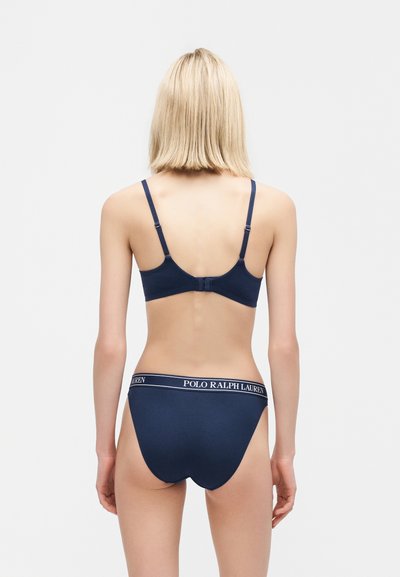 Ξανθιά γυναίκα με θέα από πίσω, φοράει μπλε σκούρο σουτιέν και αντίστοιχο εσώρουχο με λωρίδα μέσης "Polo Ralph Lauren" σε απλό λευκό φόντο.