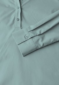 Camicia azzurra chiara con tessuto morbido e liscio. Presenta una patta con bottoni e un polsino a manica piegata.