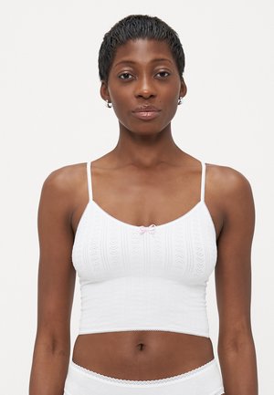 Vrouw met kort haar in een witte, gepatternde, korte camisole en bijpassend wit ondergoed tegen een effen achtergrond.