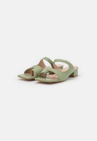 Sandali scivolati in pelle verde con punta aperta, cinturini curvi e tacco quadrato basso. Presentano una texture liscia e un design minimalista.