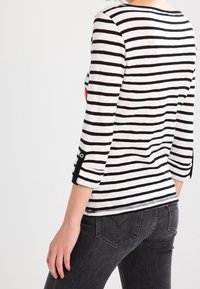 Czarna i biała, pasiasta koszula z długim rękawem i rękawami o długości trzech czwarte. Akcenty w postaci guziczków po bokach oraz zaokrąglony dół. Noszona z ciemnymi dżinsami.