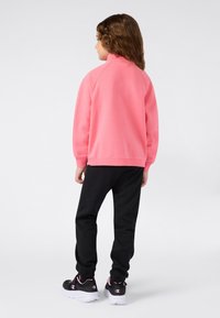 Bambino con capelli castani mossi si trova di spalle indossando una felpa rosa brillante, pantaloni della tuta neri e sneakers nere con suole bianche.