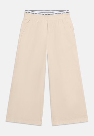 Bézs, széles szárú nadrág elasztikus derékrésszel, melyen fekete "GUESS" logó ismétlődik fehér szalagban, oldalzsebekkel és sima anyagból.