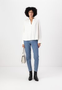Hvit langermet bluse med V-hals, kombinert med lyseblå jeans og svarte høye støvler. Modell som holder en liten grå håndveske.