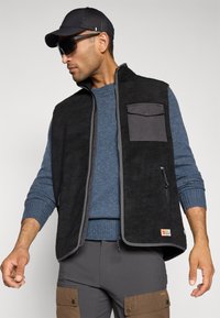 Bărbat îmbrăcat cu un vestă din fleece neagră cu fermoar și buzunar pe piept peste un pulover albastru, pantaloni gri cu buzunare maronii, o șapcă neagră și ochelari de soare.