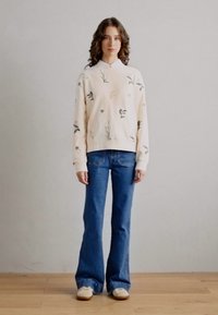 Lys rosa genser med blomstermotiv, kombinert med blå flagrende jeans og beige joggesko, på en modell som står mot en nøytral bakgrunn.