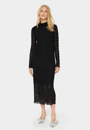 Black long-sleeve dress with a textured, diamond-patterned knit. Mid-calf length with a high neckline. Model wears silver shoes.