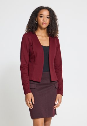 Vrouw met krullend haar, die een bordeauxkleurige blazer draagt, een bijpassende rok met zakken en een zwarte top, staand tegen een eenvoudige lichte achtergrond.