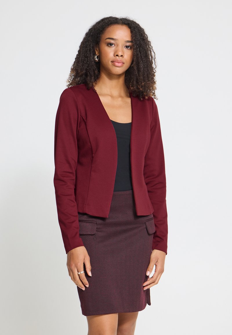 Femme aux cheveux bouclés portant un blazer bordeaux, une jupe assortie avec des poches et un top noir, debout devant un fond clair uni.
