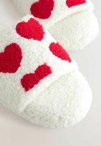 Des chaussons blancs en peluche présentent des accents en forme de cœur rouge, avec un matériau texturé et doux et un design à bout ouvert, offrant un aspect cosy.