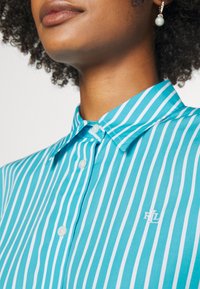 Randig turkos skjorta med knäppkrage, prydd med smala vita ränder och en broderad logotyp på vänster bröst.