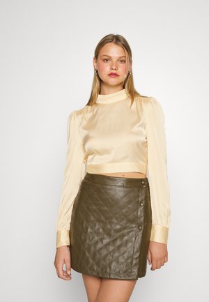 Top cropped em satin creme com decote alto e mangas bufantes, combinado com uma saia mini de couro oliva texturizada com quilt diagonal e botões.