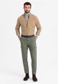 Tan overhemd met knopen, grijze coltrui, groene cargobroek, bruine riem en donkerbruine enkellaarzen. Casual fit met meerdere zakken.