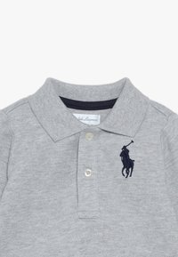 Šedé polo triko vyrobené z texturované tkaniny, s tří knoflíkovou légou, námořnickým límcem a vyšitým tmavě modrým logem na hrudi.