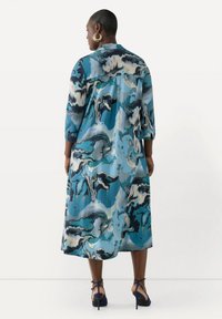 Un lungo abito blu e verde con un motivo a fantasia, dalla silhouette fluida, con maniche a tre quarti e uno spacco posteriore, abbinato a tacchi blu navy.